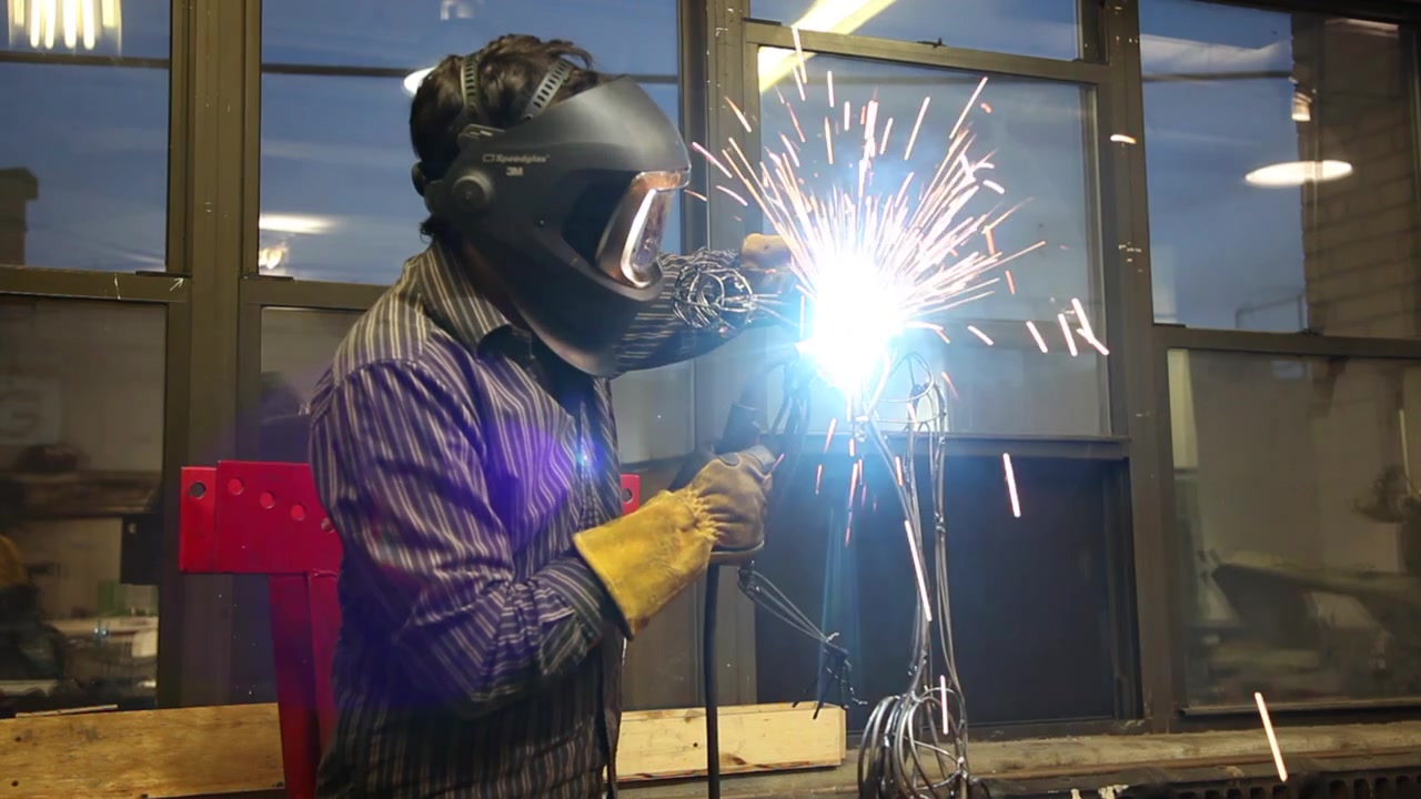 Sparks and smoke are seen as a man wearing a welding helmet and gloves uses an arc welder on a metal armature.