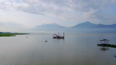 Aerial-view-of-dredger-boat-on-lake-with-crane-at-sunrise---Beautiful-mountains-and-clouds-in-backdrop