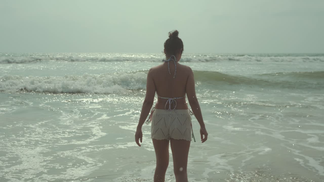 Back view of woman standing at shoreline with ocean waves and sunlight calm coastal travel scene