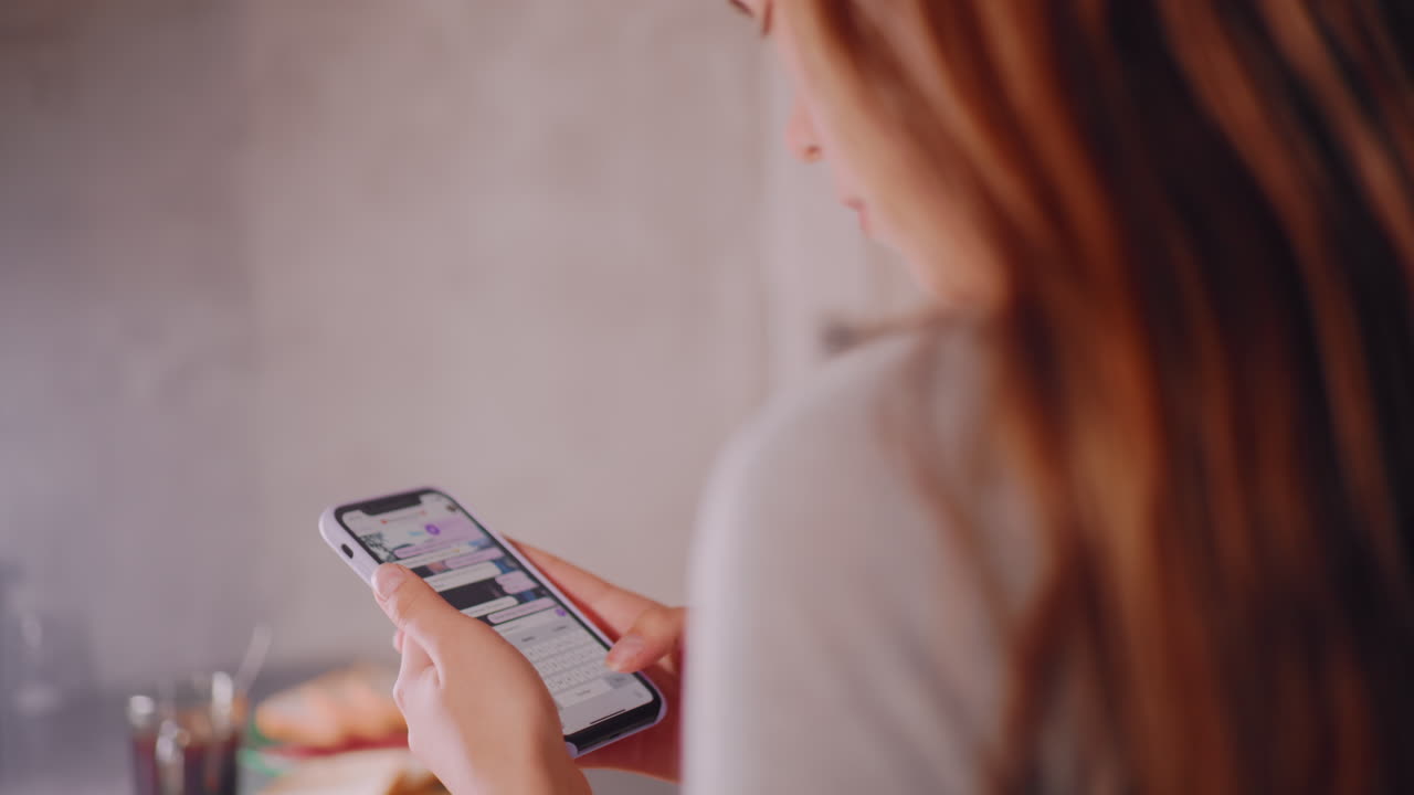 Close up of adult holding smartphone while reading chat messages in kitchen, sliced cucumber and bread on cutting board with coffee cup in blurred background