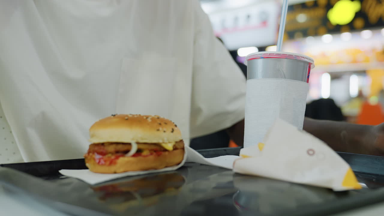 Close up person grabbing disposable drink cup beside sesame seed burger with onion ketchup patty and wrapped fries on food tray inside modern bright restaurant with bokeh background