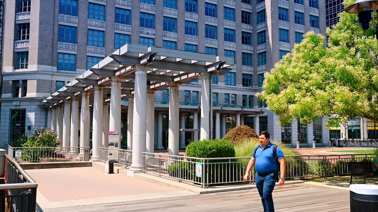 Modern urban environment with some plants. Obese Caucasian man walks by yawning. Sunny day in Jersey, USA