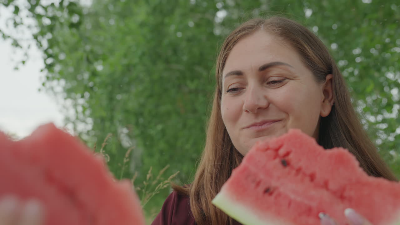 Mulher comendo melancia ao ar livre, mãe brincalhona pegando fatia oferecida pela criança, close-up na polpa vermelha e suculenta e nas sementes, árvore frondosa ao fundo, luz do dia quente, expressão sorridente, mãos e manta de piquenique