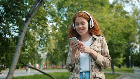 pretty redhead teenager in headphones walking outdoors in park using smartphone