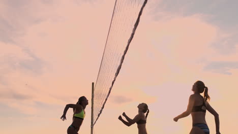 slow motion low angle close up lens flare: young female volleyball players pass and spike the ball over the net on a sunny summer evening. fit caucasian girls playing beach volleyball at sunset.