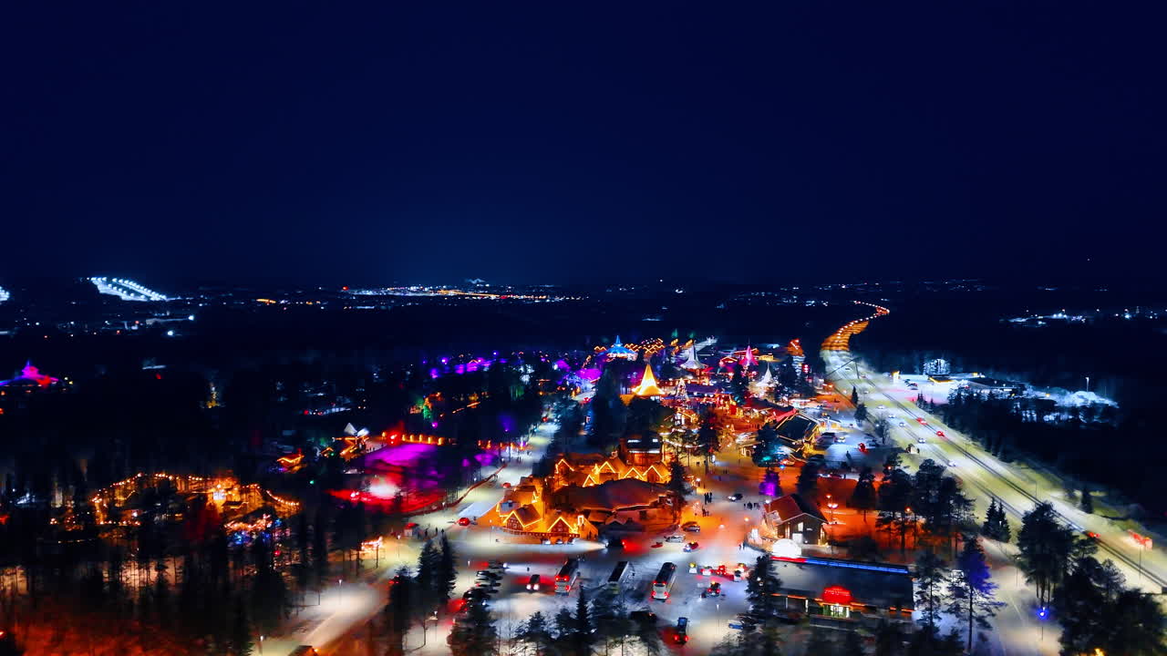 Luftaufnahme einer verschneiten Straße und eines beleuchteten Dorfes bei Nacht. Eine lange Straße führt in einer Winternacht zu einem hell erleuchteten arktischen Dorf