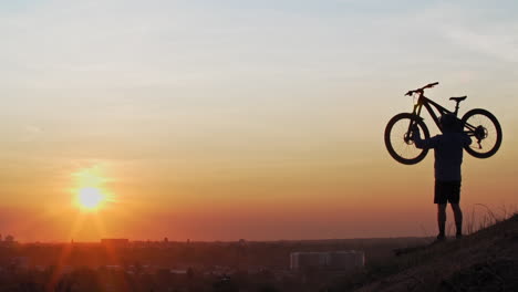 un ciclista de montaña masculino, con vista al horizonte de la ciudad en una colina, levanta su bicicleta sobre su cabeza mientras el sol bajo perfila su forma