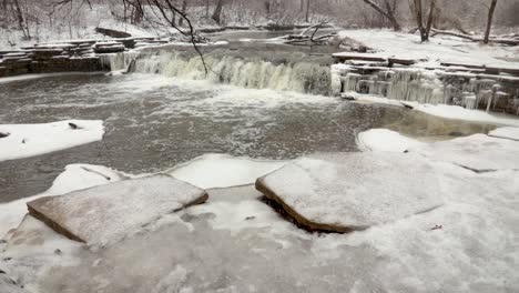 An-up-vertical-panning-shot-of-frozen-water-in-nature,-revealing-a-small-river-waterfall-in-winter-during-snowy-cloudy-afternoon