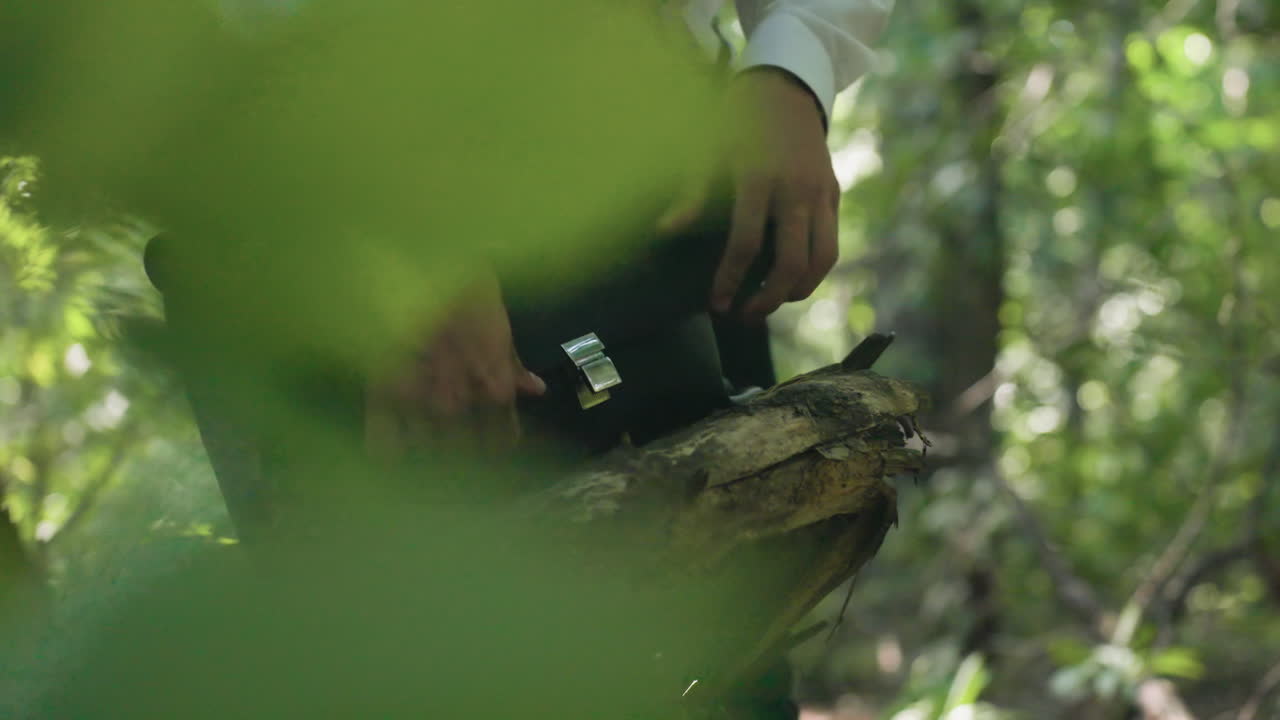 Close-up view of man in white shirt placing white coat inside black bag resting on fallen tree in forest, surrounded by greenery and natural sunlight creating woodland atmosphere