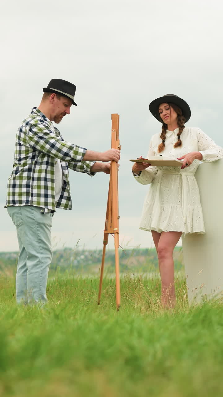 пара стоит в травяном поле, когда они готовятся рисовать на открытом воздухе. мужчина, одетый в каркасную рубашку и черную шляпу, устанавливает шезлонг, в то время как женщина, одетая в белое платье и