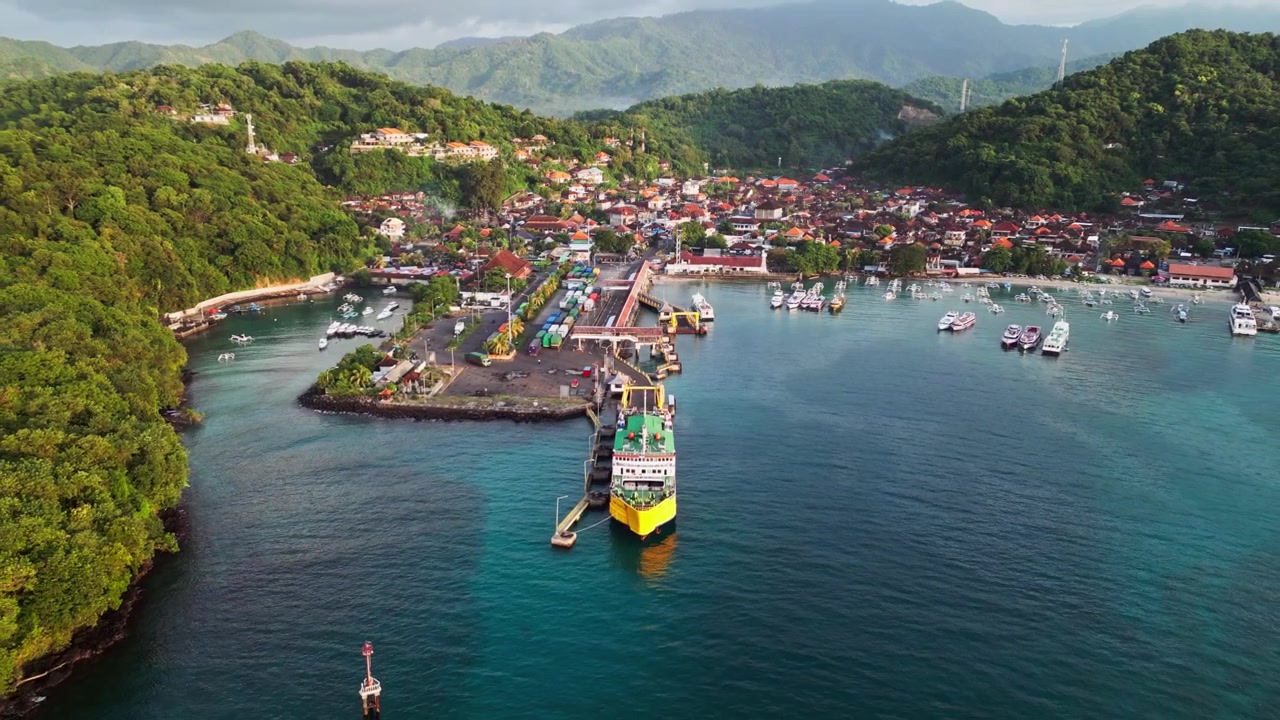 Porto costiero tropicale con terminal dei traghetti, marina e villaggio sul lungomare circondato da lussureggianti colline verdi e un oceano blu cristallino, riprese aeree con drone in allontanamento