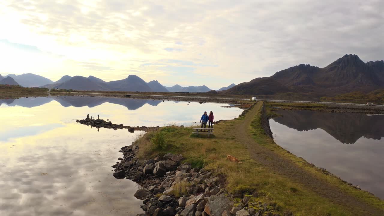 A couple standing by the water, with mountains reflecting in the calm water at Vestarelen, Norway