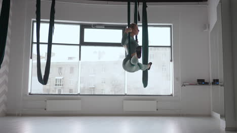 a young beautiful woman performs exercises on a hammock from aero yoga. a woman in the afternoon in front of the mirror in the hall performs stretching alone