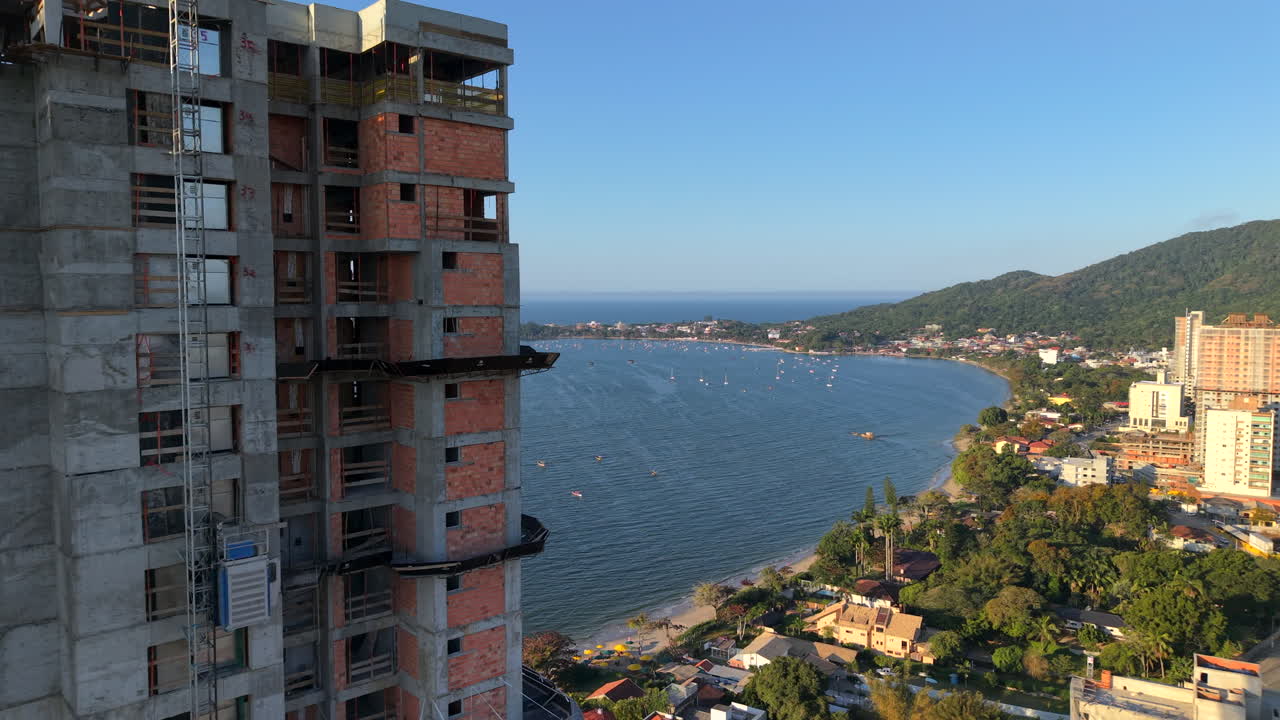 Drone desliza em direção a um arranha-céu parcialmente construído com um elevador de construção externo em Penha, Santa Catarina, Brasil, antes de revelar a baía atlântica, o litoral e as colinas arborizadas ao fundo.