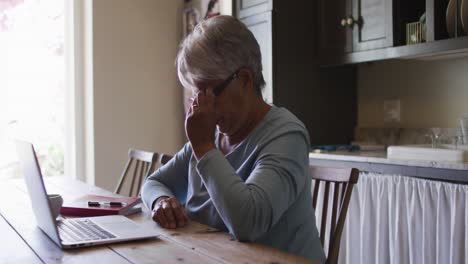 senior mixed race woman using laptop in kitchen removing glasses