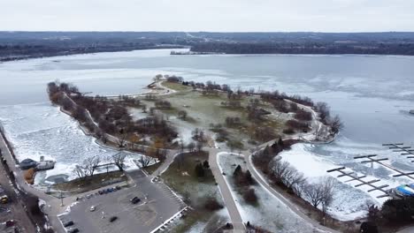 Drone-circling-over-snowy-park-next-to-a-frozen-lake-in-Hamilton