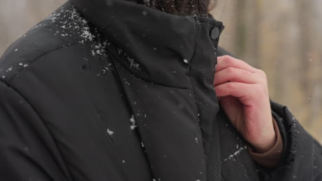 close-up of person adjusting black winter jacket strap, snowflakes visible on fabric and hair, with blurred autumnal trees in a snow-covered park background