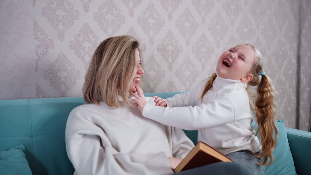 Smiling mother and daughter sitting on blue sofa at home, bonding together with joy, little girl touching mother s face playfully, family time in cozy living room, happy parent child relationship
