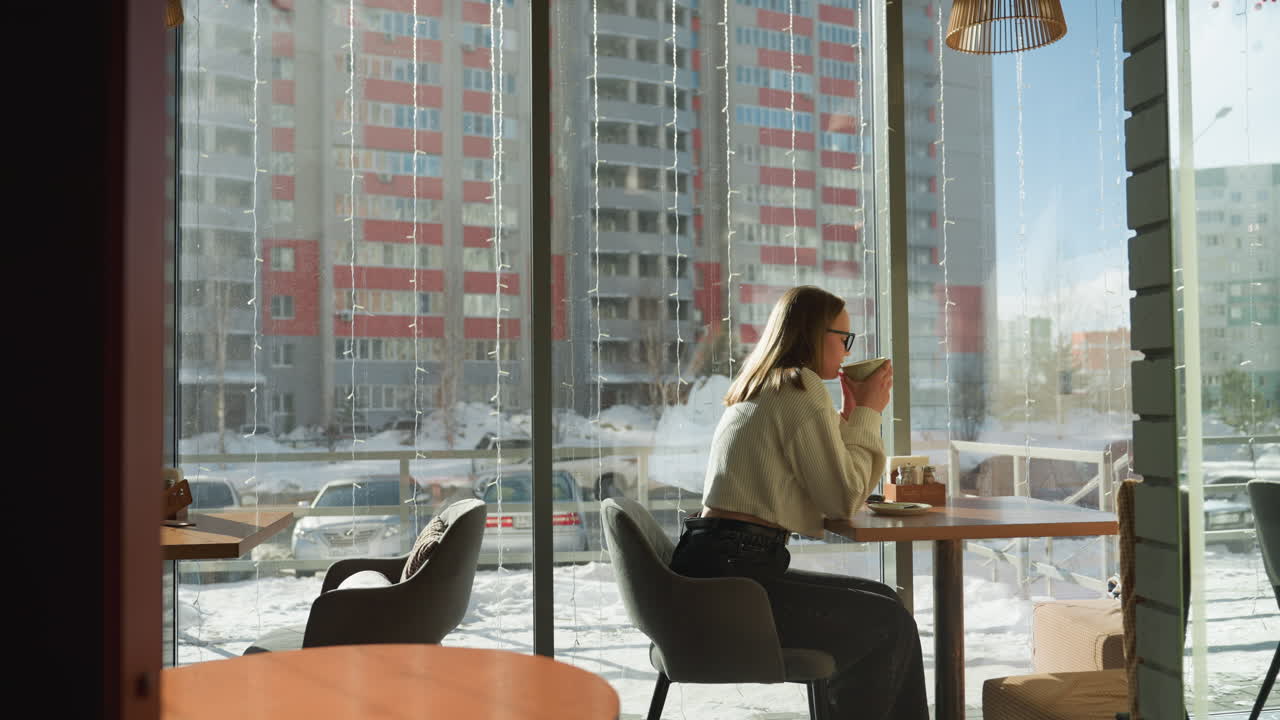 Framed interior view through wooden bars showing woman seated near window drinking tea, snowy urban background features parked cars and tall apartment buildings under clear daylight sky
