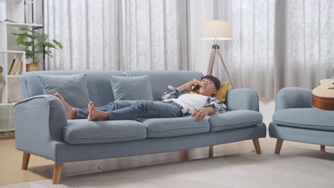 full body of asian teen boy talking on smartphone while lying on sofa in the living room at home