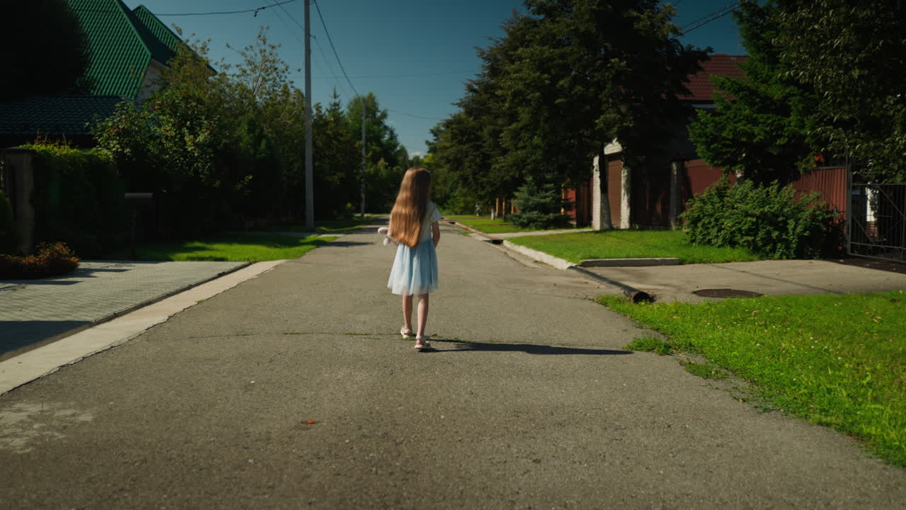Back view of young girl in blue dress walking alone along cracked residential road during sunny day with long shadow trailing behind, surrounded by green grass, trees, houses, and utility poles