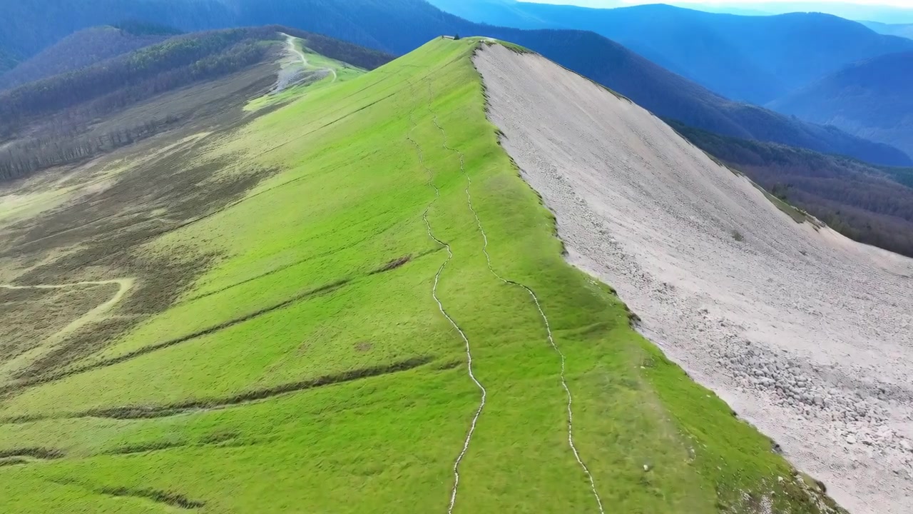 Una vista aerea simile a quella di un drone mostra una cresta montuosa verde con un sentiero roccioso grigio lungo il pendio. Un sentiero escursionistico stretto corre vicino alla cresta mentre le colline vicine sfumano in montagne blu stratificate nella morbida luce del giorno, creando un drammatico senso di profondità e natura selvaggia