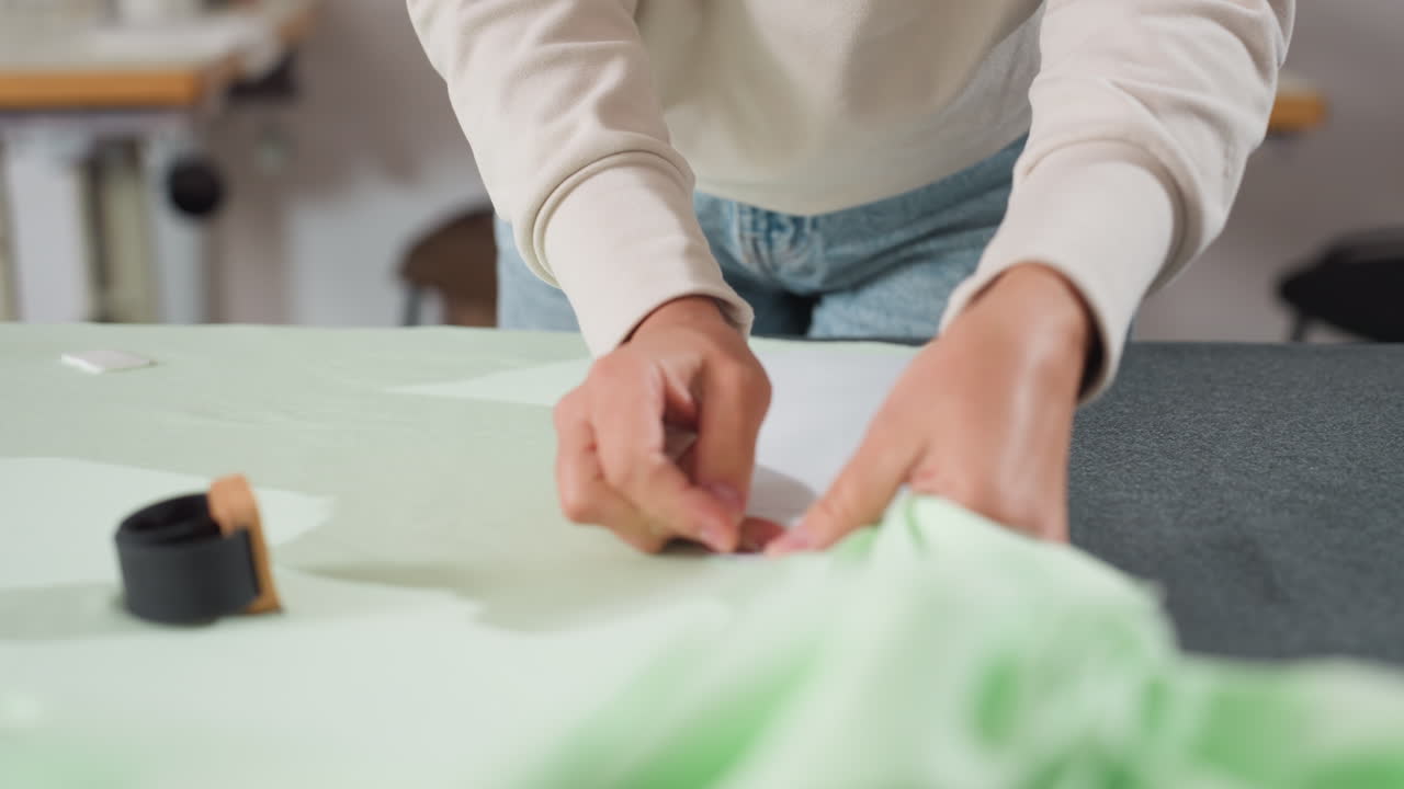 Designer carefully fixes paper cutting pattern onto spread light green fabric using pin, focusing on securing template on textile surface during garment production preparation in sewing workspace