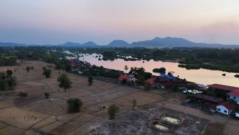 a tranquil riverside scene in don det, laos during sunset, with patchwork fields, scattered trees, houses, and distant mountains reflecting on the calm water of the mekong river