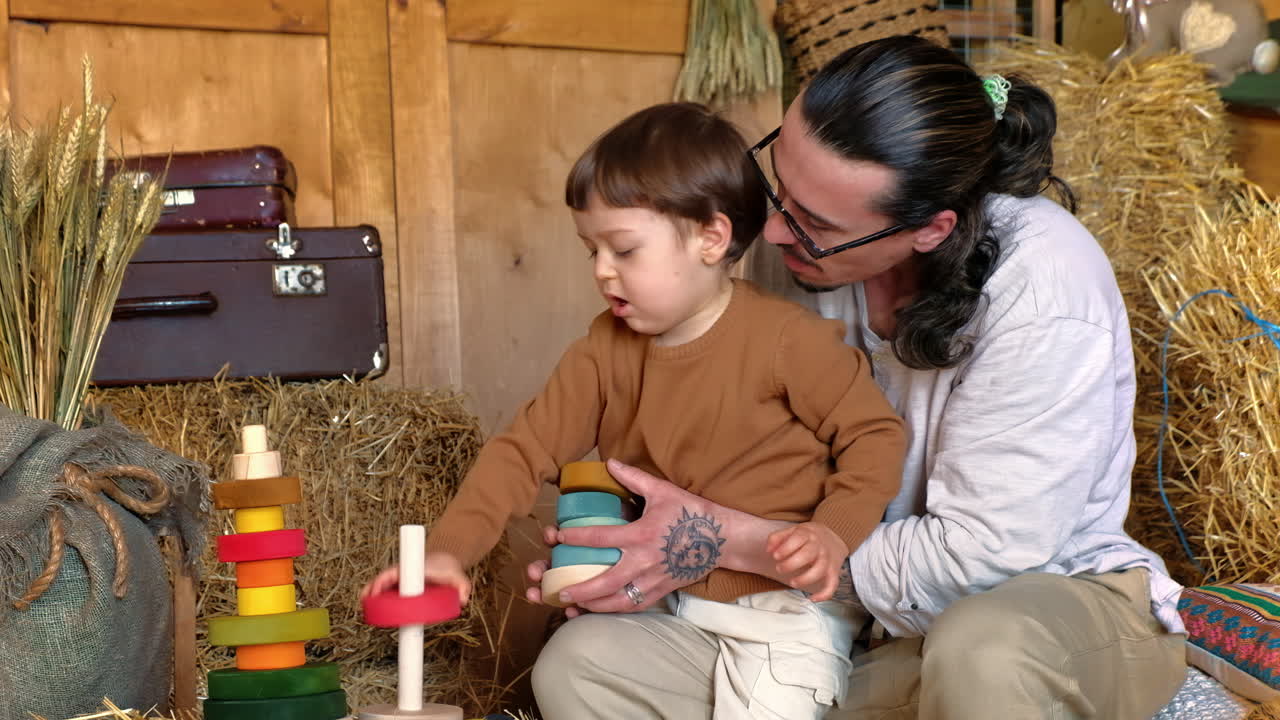 Man playing with child with colourful wooden toys in a barn, near square hay bales