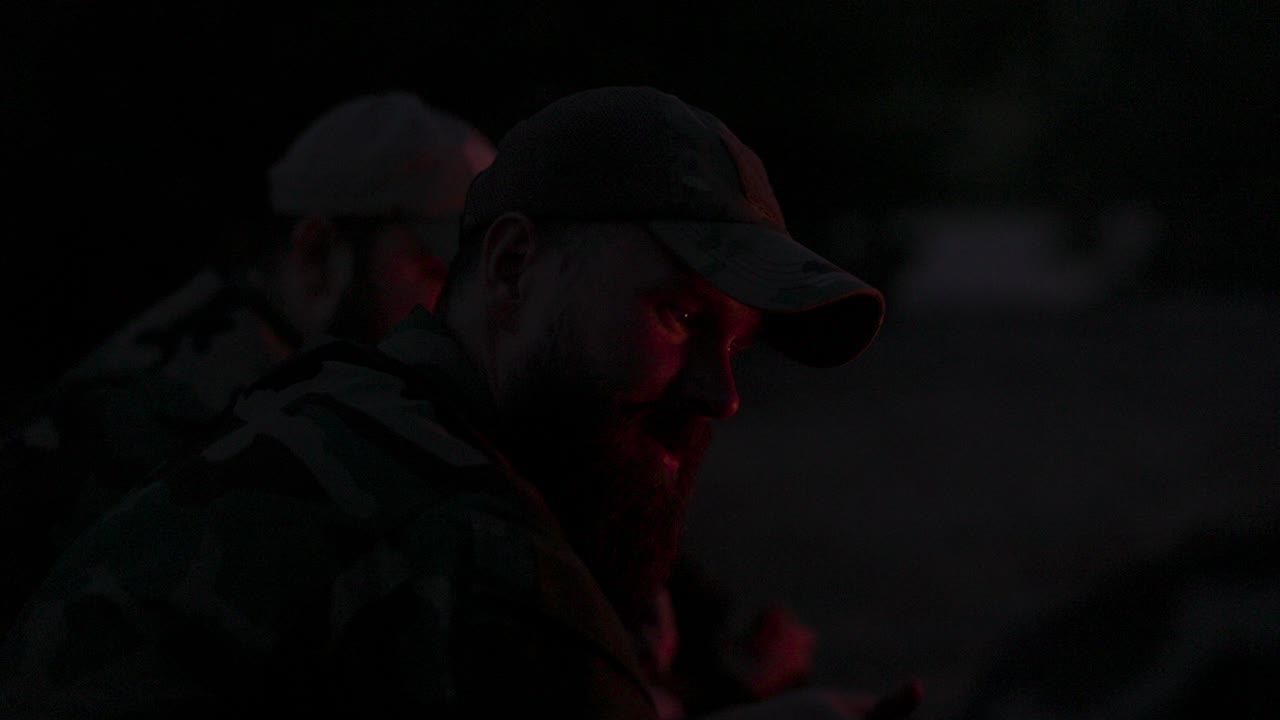 primer plano de la cara de un hombre barbudo en ropa de camuflaje y gorra por la noche iluminado por las llamas de la fogata, cuenta una historia aterradora a los amigos, confía sus problemas, campamento nocturno, caza