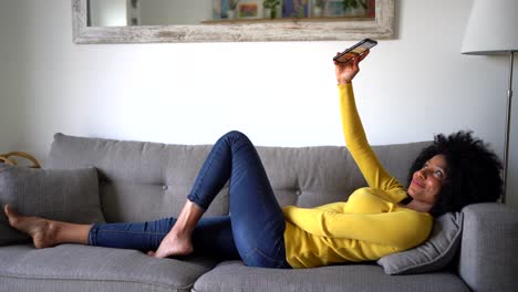 Positive black woman taking selfie with smartphone on sofa