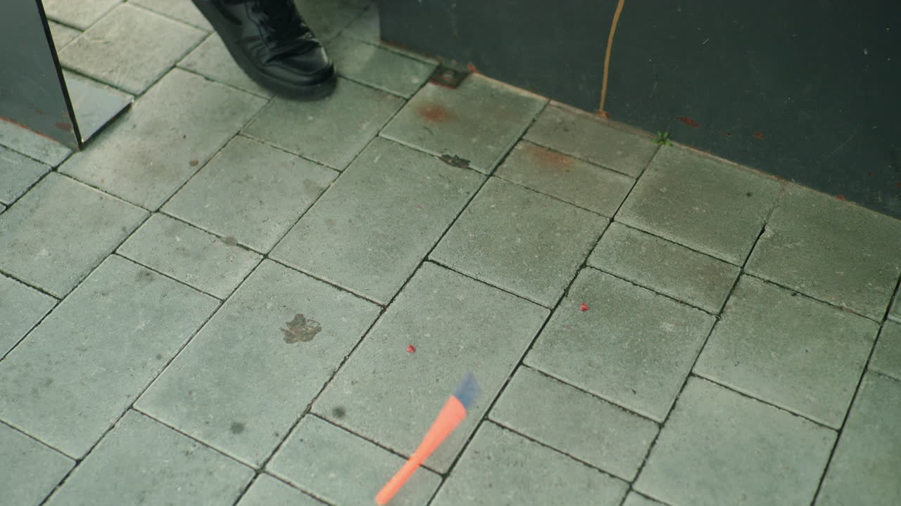 Close up view of pen falling to ground near person in black shoe on outdoor tiled pavement, capturing moment of frustration or accident with focus on object in motion and urban environment