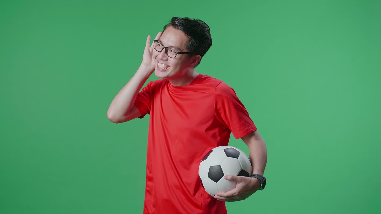 녹색 화면 배경에서 축구를 응원하는 동안 듣기 위해 공을 는 귀를 가진 아시아 남자의 측면 시각