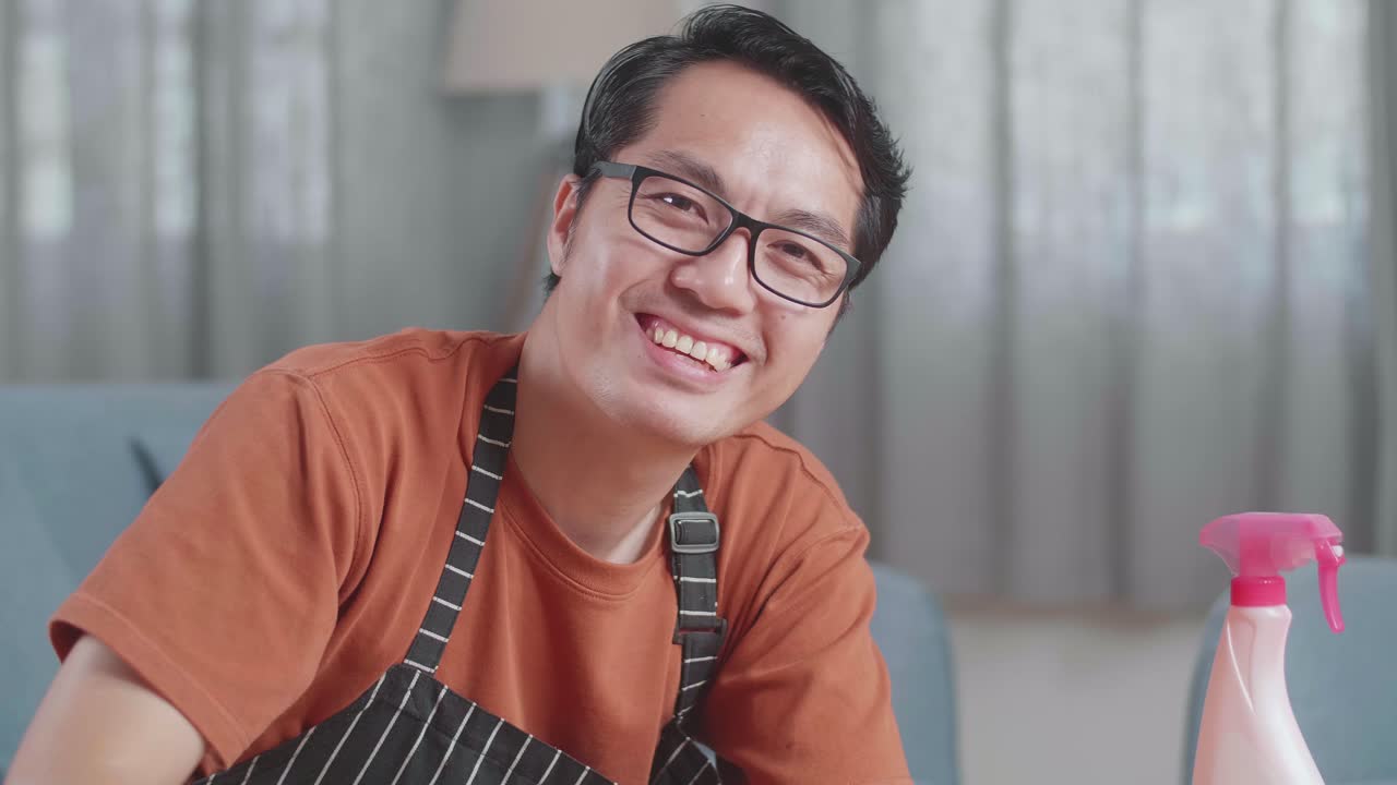 primer plano de una ama de casa masculina asiática con un delantal sonriendo y mostrando los pulgares hacia arriba gesto a la cámara mientras limpia la mesa por el spray en casa