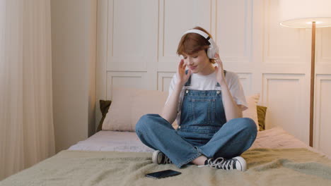 front view of girl sitting on bed wearing headphones and playing music from her smartphone