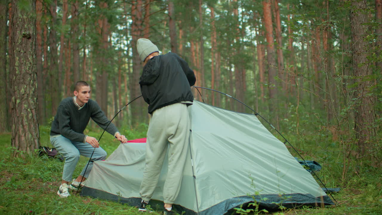 young man and woman in sweater and hoodie work together to set up camping tent in forest as one kneels holding tent edge while other stands securing pole surrounded by trees and outdoor greenery