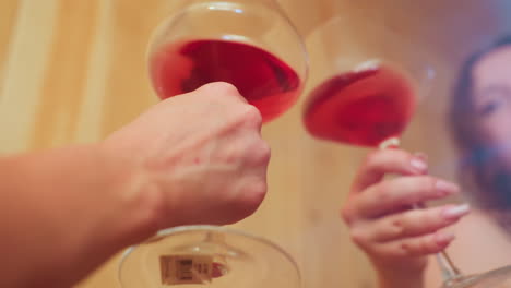 close up hands holding wine glasses mid-toast in warm intimate setting with wooden wall background, focus on ruby red liquid swirling gently
