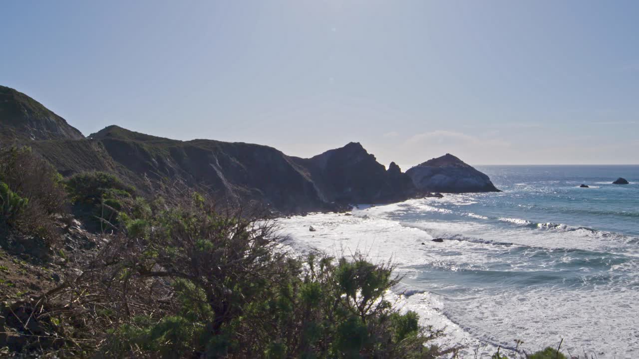 Ocean waves roll into a rugged coastline with jagged peaks and lush foreground brush under clear skies, showcasing the raw beauty of the Pacific coast