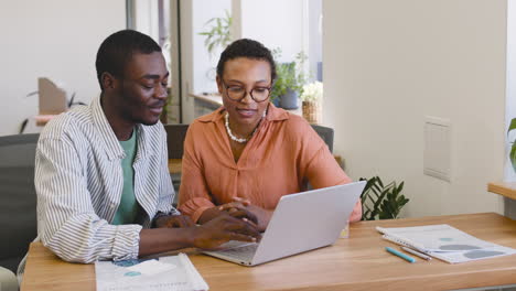 Junger-Arbeiter,-Der-Mit-Einem-Laptop-Arbeitet,-Der-An-Seinem-Schreibtisch-Sitzt,-Während-Die-Geschäftsfrau-Mit-Ihm-Spricht,-Während-Er-Am-Schreibtisch-Sitzt,-Während-Sie-Auf-Laptop-1-Schauen