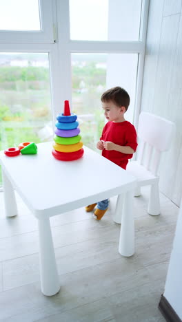 adorable caucasian kid plays with a pyramid sitting at desk. cheerful toddler builds the toy and learns the colors and sizes. vertical video.