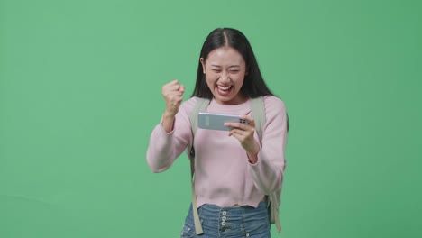 행복한 아시아 여성 학생이 모바일 게임을 하고 녹색 스크린 스튜디오에서 는 동안 승리합니다.