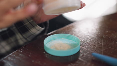 close-up view of young lady's hand tapping powder container to pour powder into lid on table, powder is spilling into lid, showcasing makeup application preparation