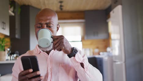 Feliz-Hombre-Afroamericano-Mayor-En-Una-Cabaña-De-Madera,-Usando-Un-Teléfono-Inteligente-Y-Tomando-Café,-En-Cámara-Lenta