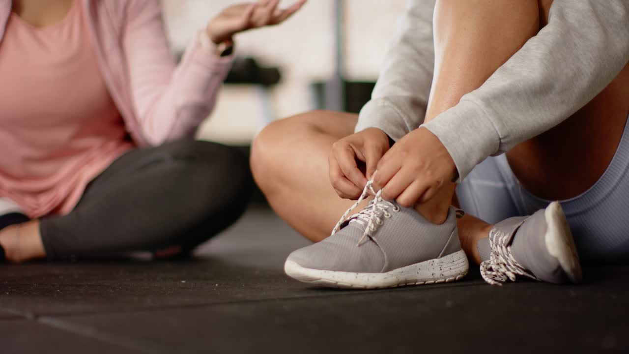 Amiche diverse che allacciano scarpe da ginnastica grigie sul pavimento della palestra seguendo i gesti dell'allenatore, copy space