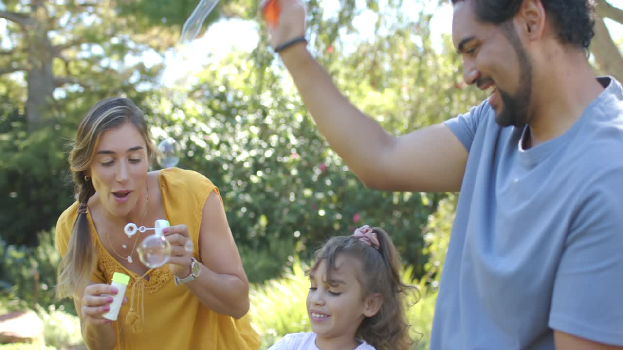 Familia de 3 en un césped soleado, mujer levantando una botella, sumergiendo una varita y soplando burbujas, enseñando a una niña.