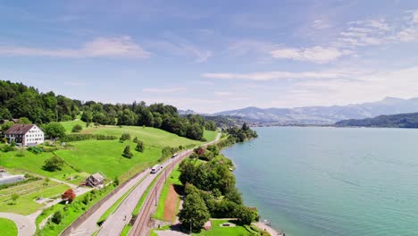 Vuelo-Lento-Hacia-Adelante-Sobre-La-Carretera-Con-Tráfico-A-Lo-Largo-Del-Lago-Superior-En-Suiza-En-Un-Hermoso-Paisaje-Con-Montañas-Y-Bosques-A-La-Luz-Del-Sol