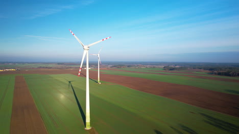 Aerial-shot-of-a-row-of-windmills-spinning-on-sunny-day-with-green-fields-and-blue-sky-in-the-background,-wind-energy-farm-generating-electricity,-green-energy-future