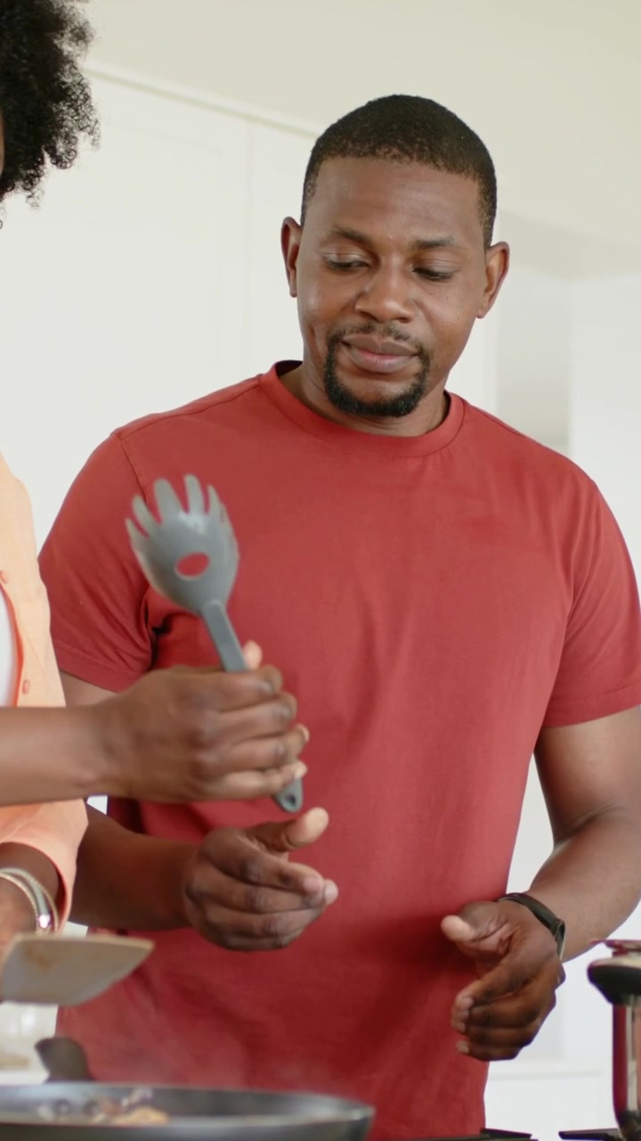 Vertical video: Seeing her stirring pan, man in red T reaching for gray server help cook in kitchen