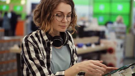 curly woman with headphones on neck standing at the counter with mobile phones in casual clothes choosing a new smartphone in a