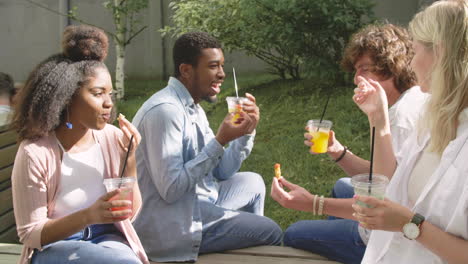 group of multiethnic friends spending time together, eating nuggets and drinking refreshing juice, sitting in a wooden bench in a park, talking to each other and having fun